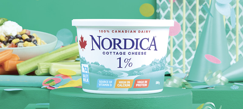 Nordica 1% cottage cheese on a stand surrounded by green party hats, wrapping paper, and confetti. A plate with a taco bowl, carrots, and celery are in the background.