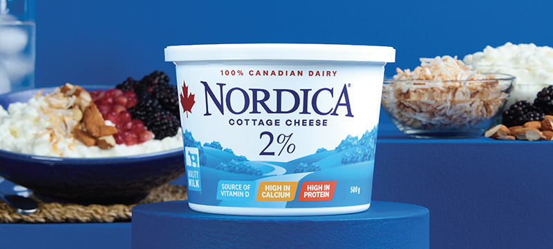 Nordica 2% cottage cheese in the foreground with a blue background. A bowl of cottage cheese with coconut, almonds, and blackberries and individual ingredients in the background.
