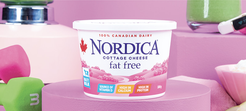 Nordica Fat Free cottage cheese on a stand surrounded by a two hand weights, a blender, a smoothie, a smart watch, apples slices, and a bowl of cottage cheese.