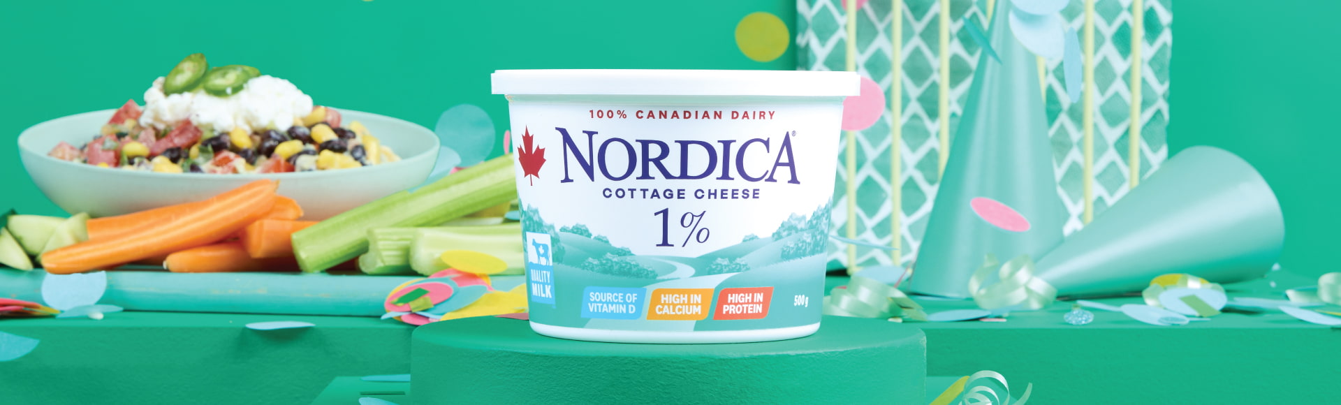 Nordica 1% cottage cheese on a stand surrounded by green party hats, wrapping paper, and confetti. A plate with a taco bowl, carrots, and celery are in the background.