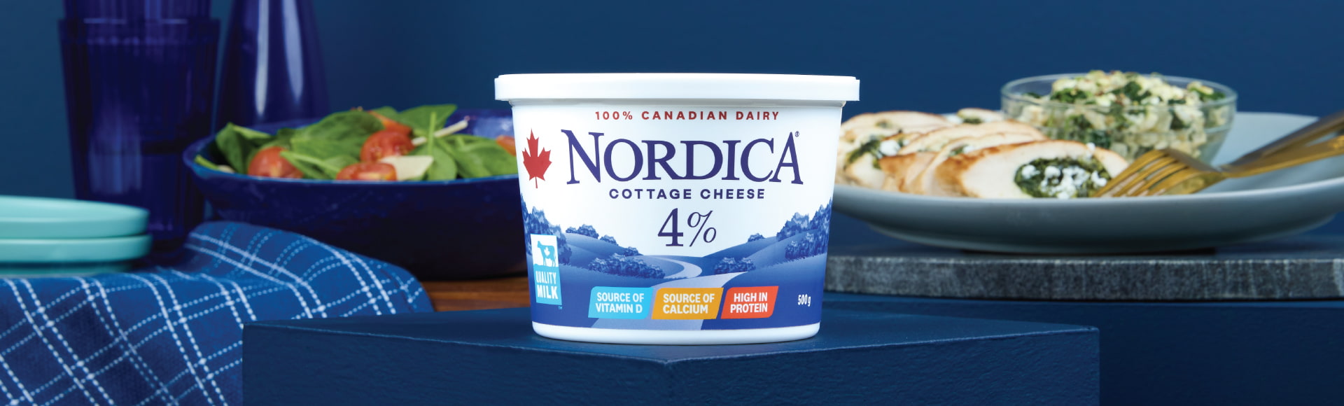 Nordica 4% cottage cheese on a stand surrounded by a plate of salad and a plate of chicken stuffed with spinach.
