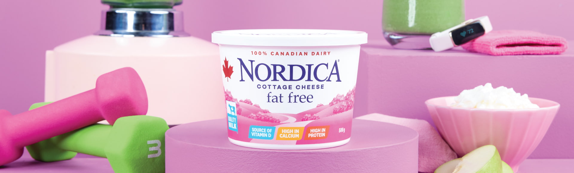Nordica Fat Free cottage cheese on a stand surrounded by a two hand weights, a blender, a smoothie, a smart watch, apples slices, and a bowl of cottage cheese.