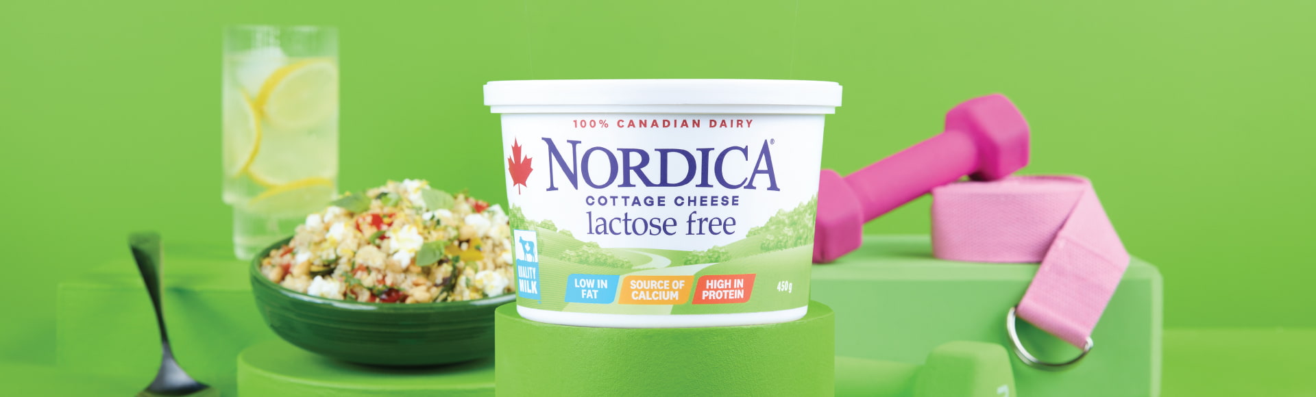 Nordica Lactose Free cottage cheese on a stand surrounded by a weight, a belt, a glass of ice water with lemon, a spoon, and a bowl of quinoa and cottage cheese.